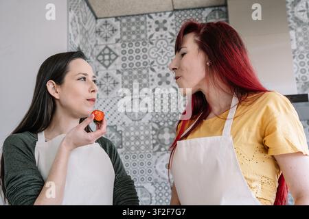 Two women in aprons tasting a strawberry in a modern kitchen Stock Photo
