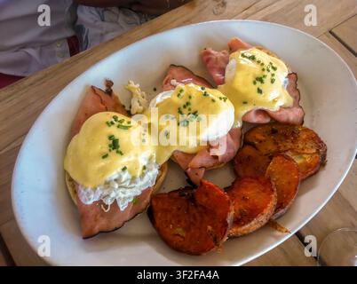 A high-angle close-up shot of a white plate serving traditional Eggs Benedict. The dish features poached eggs served over ham slices on English muffin Stock Photo