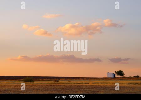 A serene late afternoon view in Alhambra, Spain, featuring a small white rural building under a glowing sunset sky with soft, illuminated clouds. Stock Photo