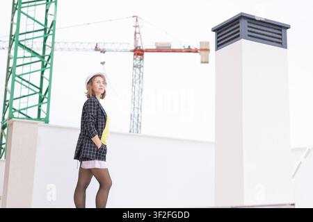 Female architect or construction engineer on a building rooftop analyzing the urban development plan, wearing a hard hat, jacket, and glasses, with ci Stock Photo
