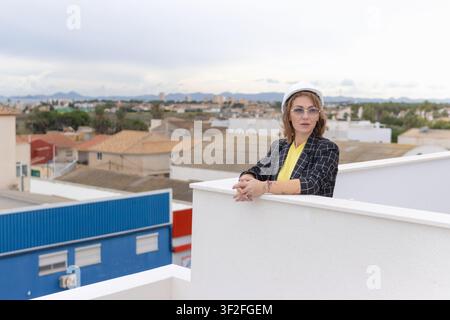 Female architect in hard hat and sunglasses standing on a rooftop, observing urban development and construction site, planning future city infrastruct Stock Photo