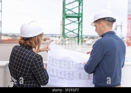 Architects wearing hard hats are collaborating on a rooftop construction site, carefully examining blueprints and planning future building phases whil Stock Photo