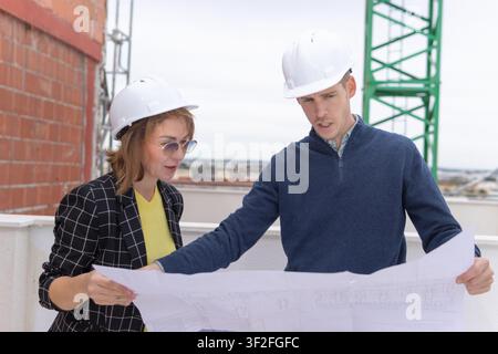 Two construction professionals, an architect and a manager, reviewing building plans on a construction site, collaborating on project development and Stock Photo
