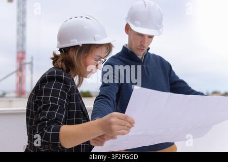 Two architects or engineers wearing safety helmets and reviewing blueprints on a construction site, collaborating and discussing building plans for a Stock Photo