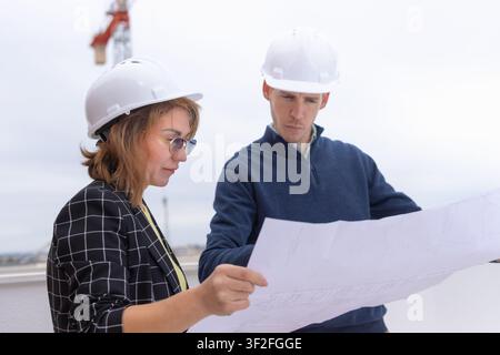 Civil engineers and architects discussing building development plans on construction site, holding blueprints, and wearing hard hats for safety in the Stock Photo