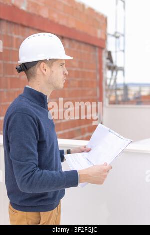 Young male engineer wearing a white hardhat and casual clothing, reviewing architectural blueprints while standing on a building under construction, f Stock Photo