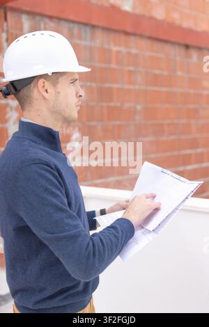 Professional engineer wearing a white hard hat and blue pullover checking architectural blueprints and construction documents on a new building site w Stock Photo