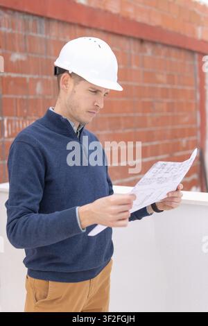 Construction professional wearing a safety hard hat, carefully examining architectural plans and blueprints on a building site, overseeing work progre Stock Photo
