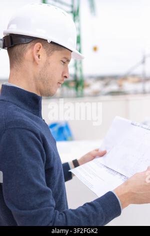 Engineer wearing a white hard hat and blue sweater carefully studying blueprints at a construction site. Ensuring meticulous project planning. Safety Stock Photo