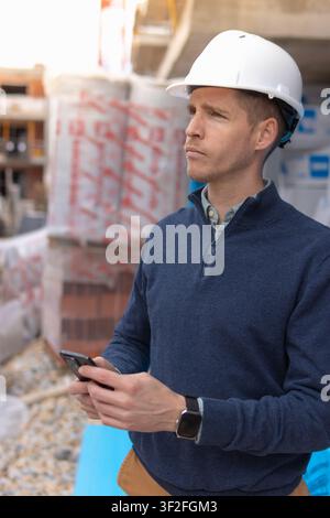 Male building project manager in a white safety hard hat and casual attire using a smartphone at a construction site, overseeing development and plann Stock Photo
