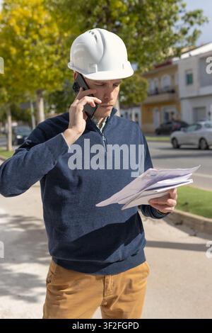 Male professional engineer or architect managing construction details, communicating on a mobile phone and reviewing blueprints while working on a pro Stock Photo