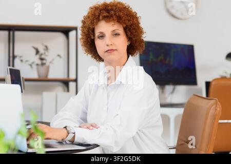 Confident businesswoman with curly red hair working on a laptop in a modern office setting, She is wearing a white shirt and a watch, with her arms cr Stock Photo