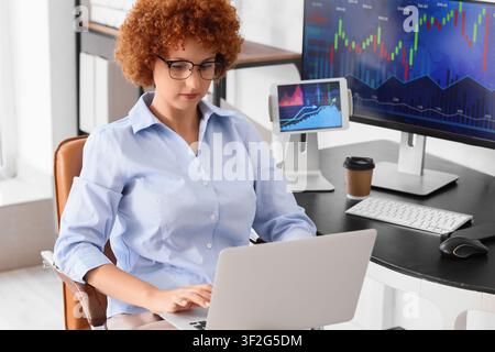Young woman with curly red hair working on a laptop at a desk with Multiple screens displaying financial charts, Multiple computer screens display fin Stock Photo