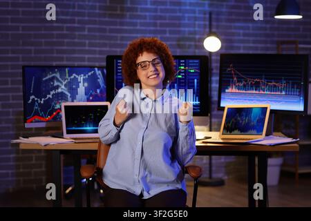 Excited young woman celebrating financial success with fists raised in front of multiple computer screens displaying stock market data at night Stock Photo