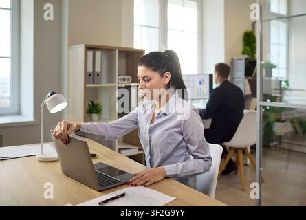 Focused businesswoman working on laptop at desk in modern office with coworker Stock Photo