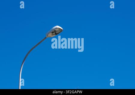 Low angle view of a modern public street light mounted on a curved metal pole. The lamppost stands isolated against a vibrant, cloudless blue sky Stock Photo