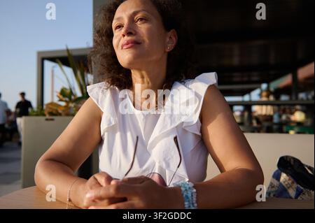 A confident woman at an outdoor cafe table bathed in warm sunlight, wearing a white dress and bracelets, looking thoughtful and relaxed while enjoying Stock Photo