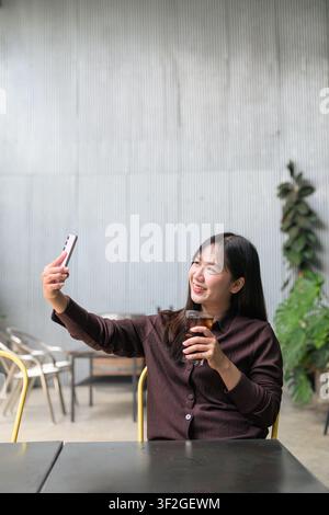 A vertical composition showing a happy young Asian woman taking a selfie with her pink smartphone at an outdoor restaurant table. She smiles warmly to Stock Photo