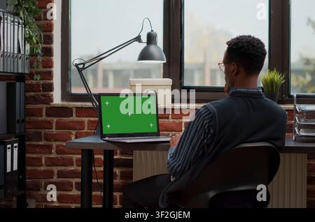 Secretary Using Laptop with Green Screen Mockup uses a laptop chroma key screen in a software development workspace, with blank copy space and isolated mockup background for editable screen. Stock Photo