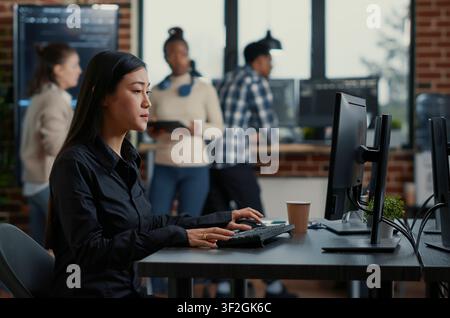 Asian software developer sits at a desk with a clipboard and starts typing on a computer keyboard while programmers analyze code on a wall screen. Stock Photo
