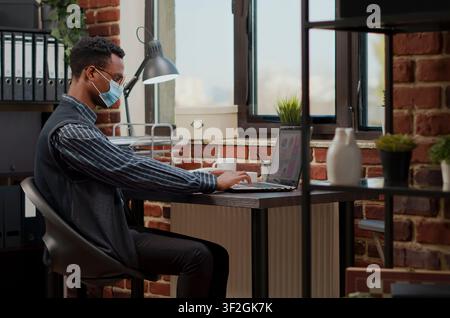 Masked businessman handles an ecommerce task at an office desk, using a laptop to plan financial statistics, sales work and company development. Stock Photo