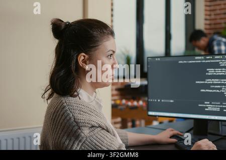 Focused software engineer writes code while viewing multiple computer screens with programming language algorithms as colleagues collaborate in the background. Stock Photo