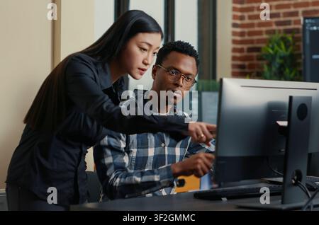 Two software developers sit at a desk with a laptop coding interface, pointing at computer screens while discussing algorithms and source code. Stock Photo