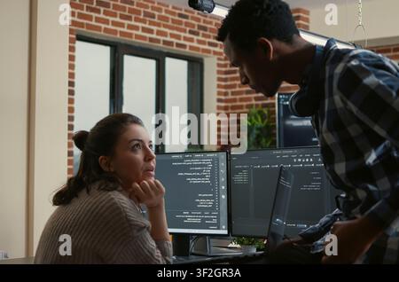 Junior developer sits on a desk and shows a laptop with source code to a senior programmer, interrupting algorithm work to ask for help fixing errors. Stock Photo