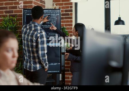 System developers analyze code on a wall screen while checking a digital tablet for errors, comparing results with a team of coders in a startup office. Stock Photo