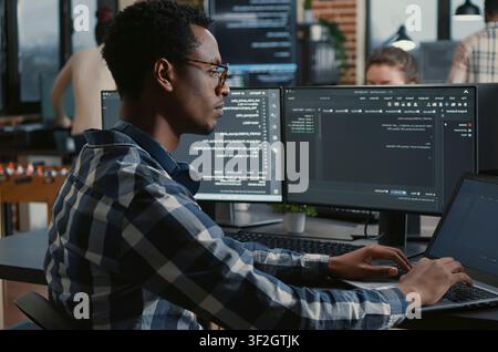 Focused programmer adjusts glasses while typing on a laptop, parsing code on multiple screens as teammates work on big data and user interface tasks in the background. Stock Photo