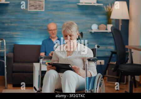 Aged woman in a wheelchair holds a digital tablet and browses the internet, using touch screen technology while managing physical disability at home. Stock Photo
