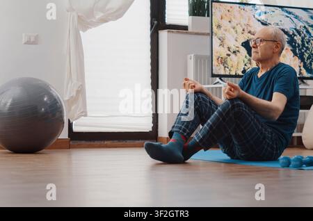 Senior man sits in lotus position on a yoga mat and meditates after exercise, using breathing, relaxation and wellness practice for active aging. Stock Photo