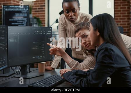 Programmer points at a computer screen to explain running code while coworkers brainstorm software ideas, collaborating on an IT group project in a freelance agency. Stock Photo