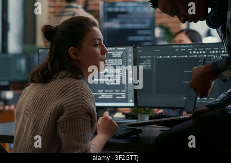 Junior programmer sits on a desk and shows laptop source code to a senior developer, asking for help with errors during algorithm work. Stock Photo