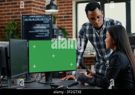 Mixed programmers analyze source code beside a green screen chroma key mockup, discussing algorithms, app development and online cloud computing in a server room. Stock Photo