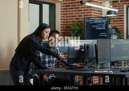 Two software developers carry a laptop with coding interface toward a desk, discussing algorithms and a group project with screens nearby. Stock Photo