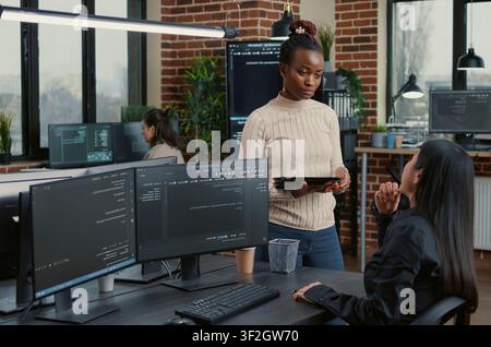 Software developer standing up holding digital tablet talking with programmer sitting at desk with computer screens showing running code around computer screens, showing developer teamwork, source. Stock Photo