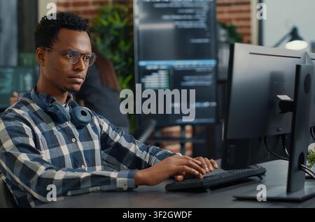 Focused programmer fixes his glasses while writing code at a desk, with teammates programming user interface work in the background. Stock Photo