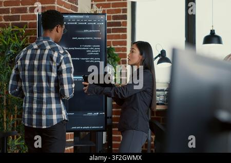 Team of coders analyzes code errors on a wall screen while using a digital tablet beside a programmer working with neural network software. Stock Photo