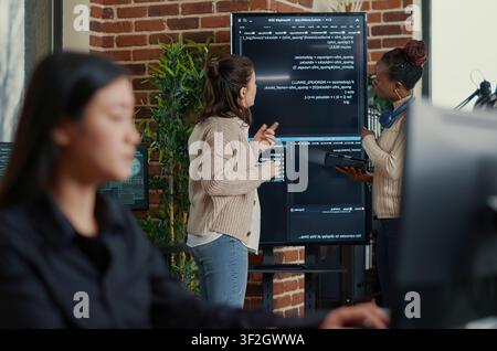 System engineers compare source code errors on a wall screen using a digital tablet while a programmer writes algorithm code during a debugging review. Stock Photo