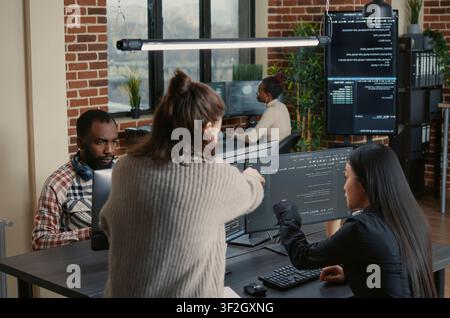 Software programmer pointing pencil at source code on computer screen explaining algorithm to coworker standing next to desk around computer screens, showing developer teamwork, source code. Stock Photo