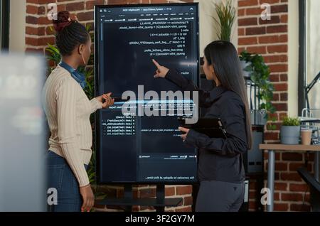 Programming team searches for code bugs as engineers use a tablet and wall display to review bugs, cloud computing and team software development in an IT office. Stock Photo