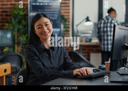 Asian software developer writes database code at a desk in a development agency, combining programming language, laptop coding and a confident technology portrait. Stock Photo