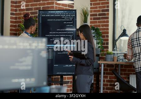 Engineer explains wall screen code errors as engineers use a tablet and wall display to review bugs, cloud computing and team software development in an IT office. Stock Photo