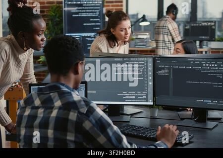 Mixed team of artificial intelligence cloud programmers talking about programming in front of multiple screens compiling code, programmers doing teamwork looking at machine learning algorithms. Stock Photo