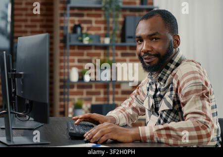 Programmer typing programming code on computer keyboard turning head and smiling while colleague walks by in background, software engineer sitting at desk writing code feeling confident. Stock Photo