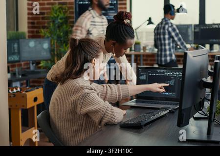 Two software coders analyzing source code looking for errors running on laptop screen while sitting at desk, team of system engineers working on cyber security innovation in busy server room. Stock Photo