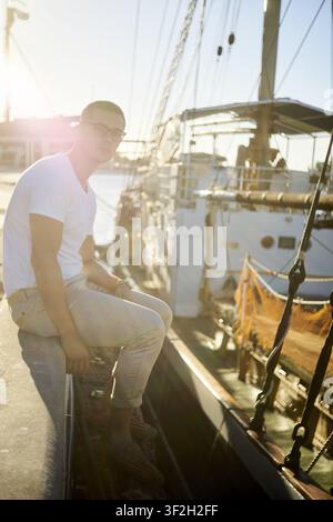 Sunlit man chilling in casual attire sitting on dock near boats with a sunny background at harbour dock, leisure hobby of sailing, boatsman, seaman, s Stock Photo