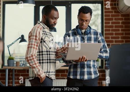 System engineers discuss algorithms while holding a laptop in an office where colleagues work on database and programming tasks for realistic commercial stock use and row-specific context. Stock Photo