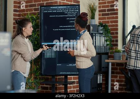 Programmer holding digital tablet analyzing code on wall screen tv while colleague is looking for errors pointing using remote control, with wall screen and African American. Stock Photo